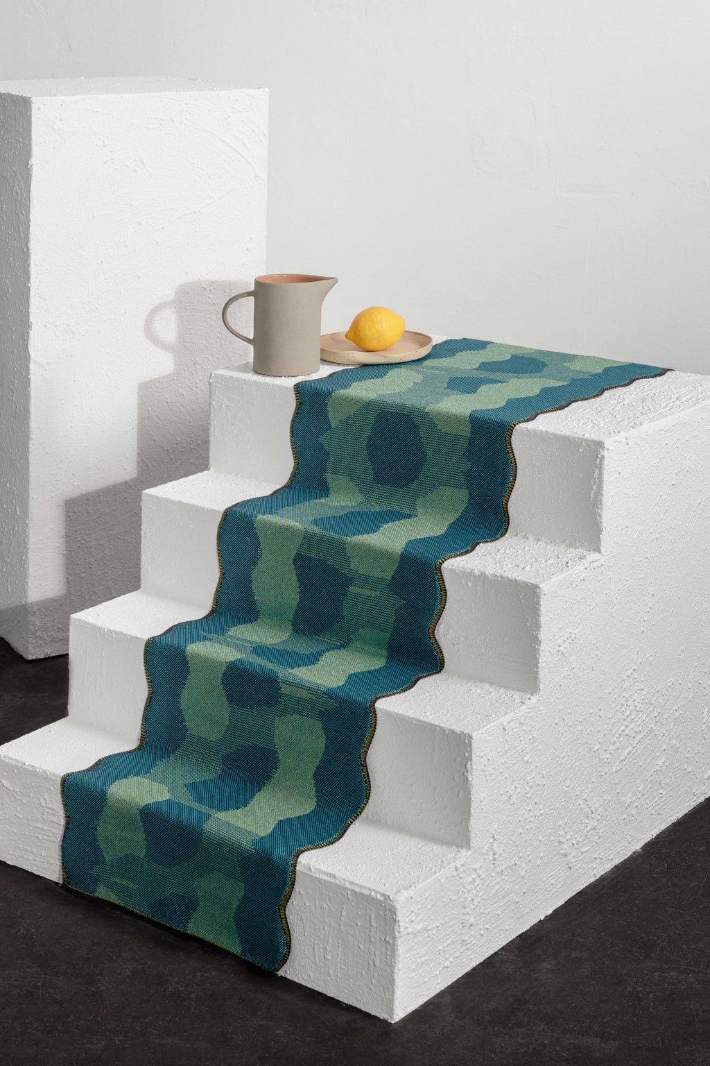 side view of green knit organic cotton and viscose table runner with abstract pattern and wavy edges on white display
