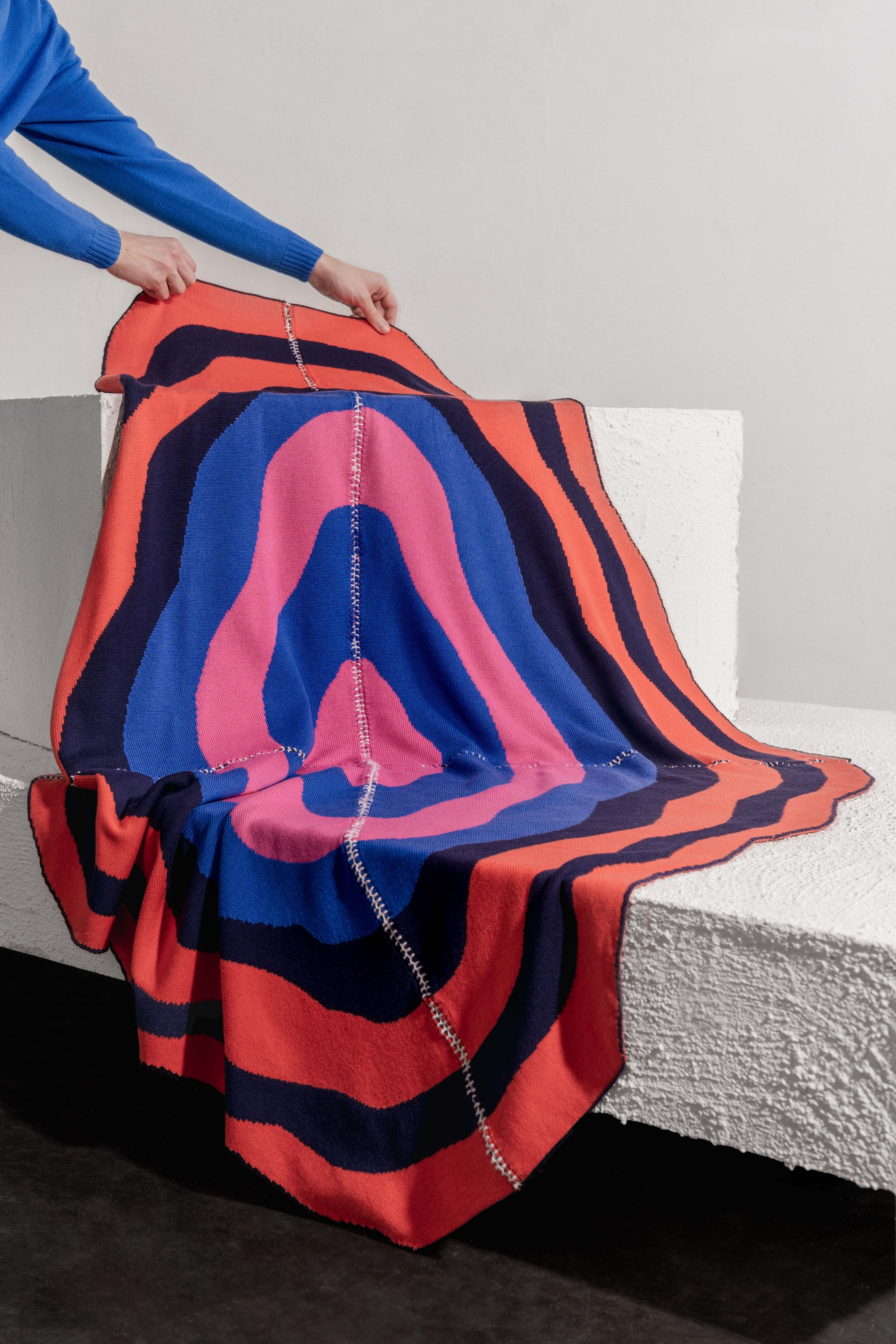 hands holding dark wavy knit wool blanket with hand-stitched seam in blue, pink, red and navy 